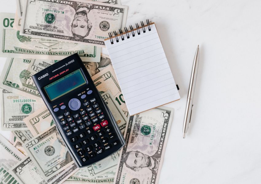 A black calculator, silver pen and small notepad sit on top of a pile of. american bills on a white desk, ready for a landlord to calculate their property rental price.
